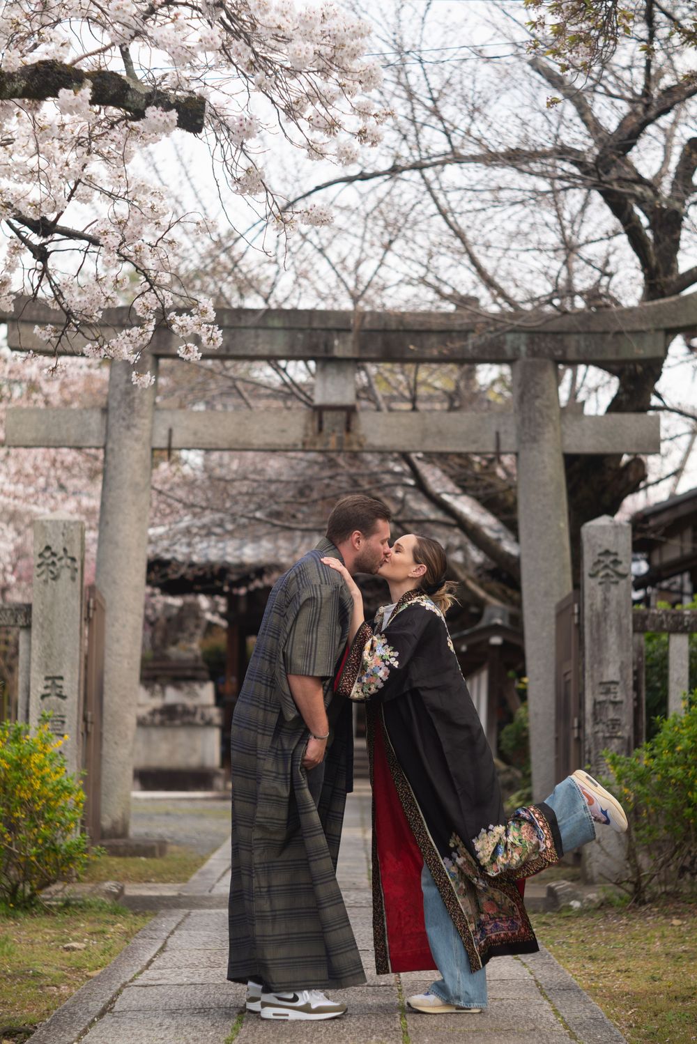 Couple in traditional Japanese kimono kissing beneath a stone torii gate at a shrine surrounded by blooming cherry blossoms.
