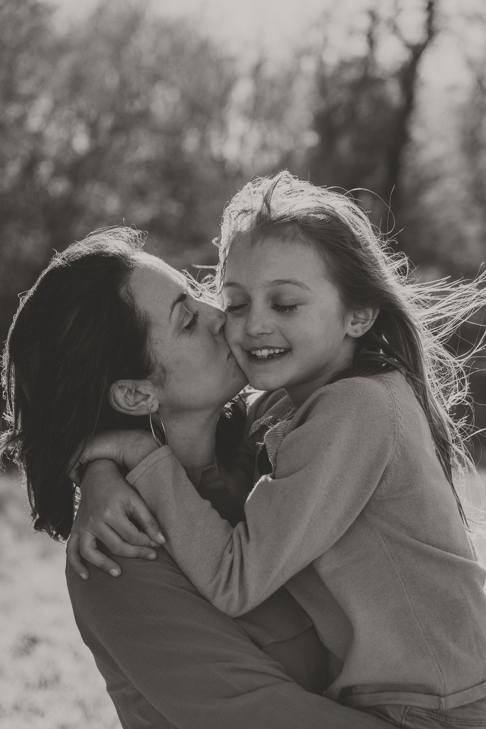 Black and white portrait of a mother kissing her smiling daughter's cheek outdoors, the girl's hair blowing in the breeze.