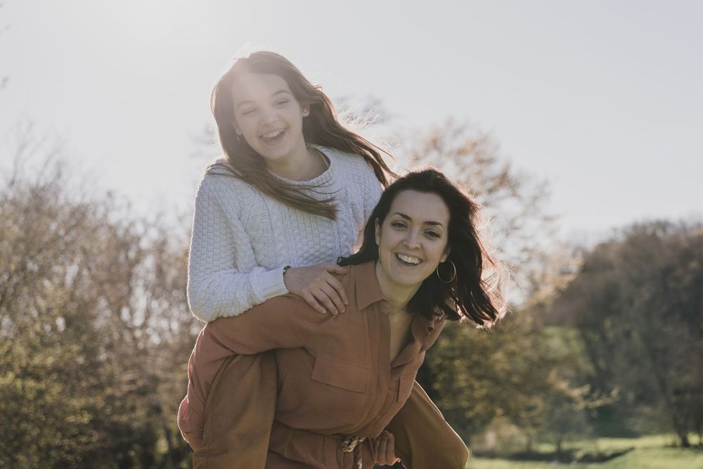 Smiling mother gives her laughing daughter a piggyback ride outdoors, with autumn trees and soft sunlight in the background.