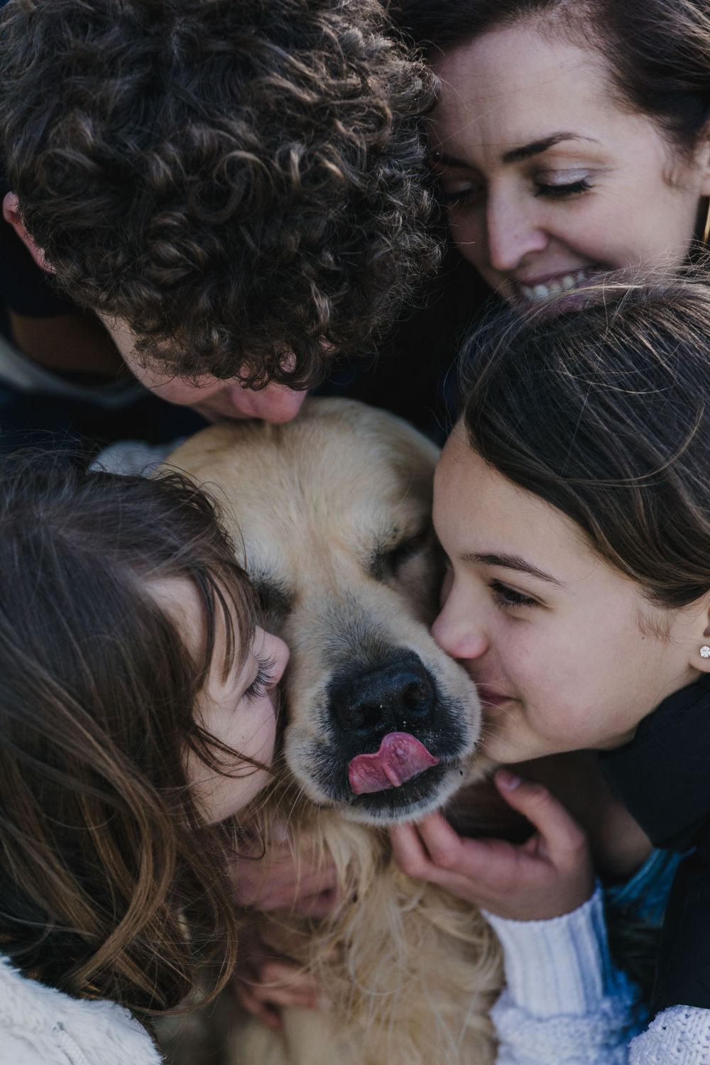Three smiling family members cuddle a happy golden retriever with its tongue out in a warm, affectionate embrace.