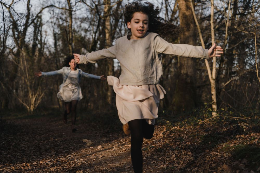 A curly-haired girl in a beige dress runs joyfully through a wooded path with her arms outstretched as another person follows.