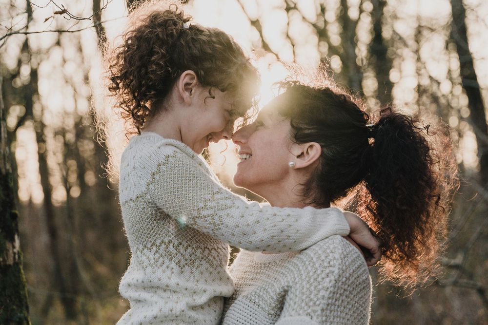A smiling mother holds her curly-haired child close, their foreheads touching in warm golden light among the trees.