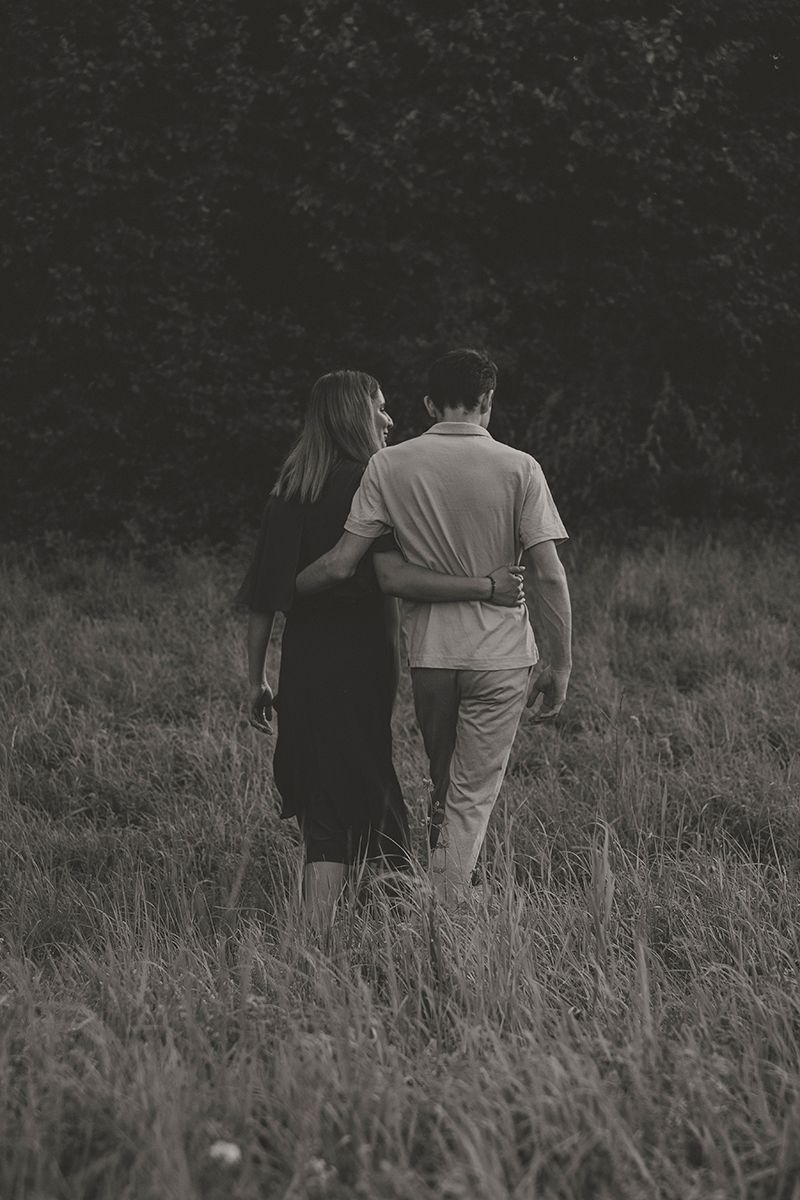 In a black and white photo, a couple walks arm in arm through tall grass toward a dark tree line.
