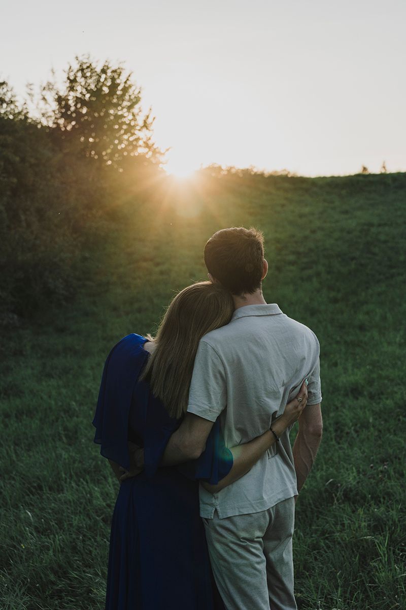 A couple embraces in a green meadow, facing a glowing sunset with sunbeams shining through the trees.