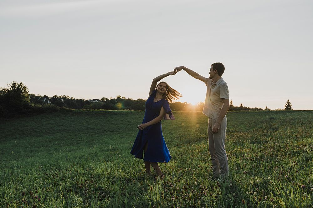 A man twirls a woman in a blue dress in a grassy field as the golden sun sets behind them.