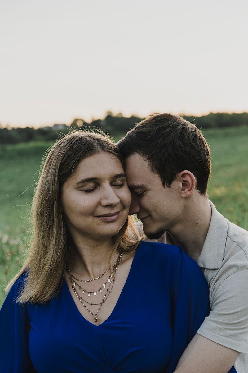 A woman in a blue dress closes her eyes and smiles as a man rests his forehead against hers in a green field.