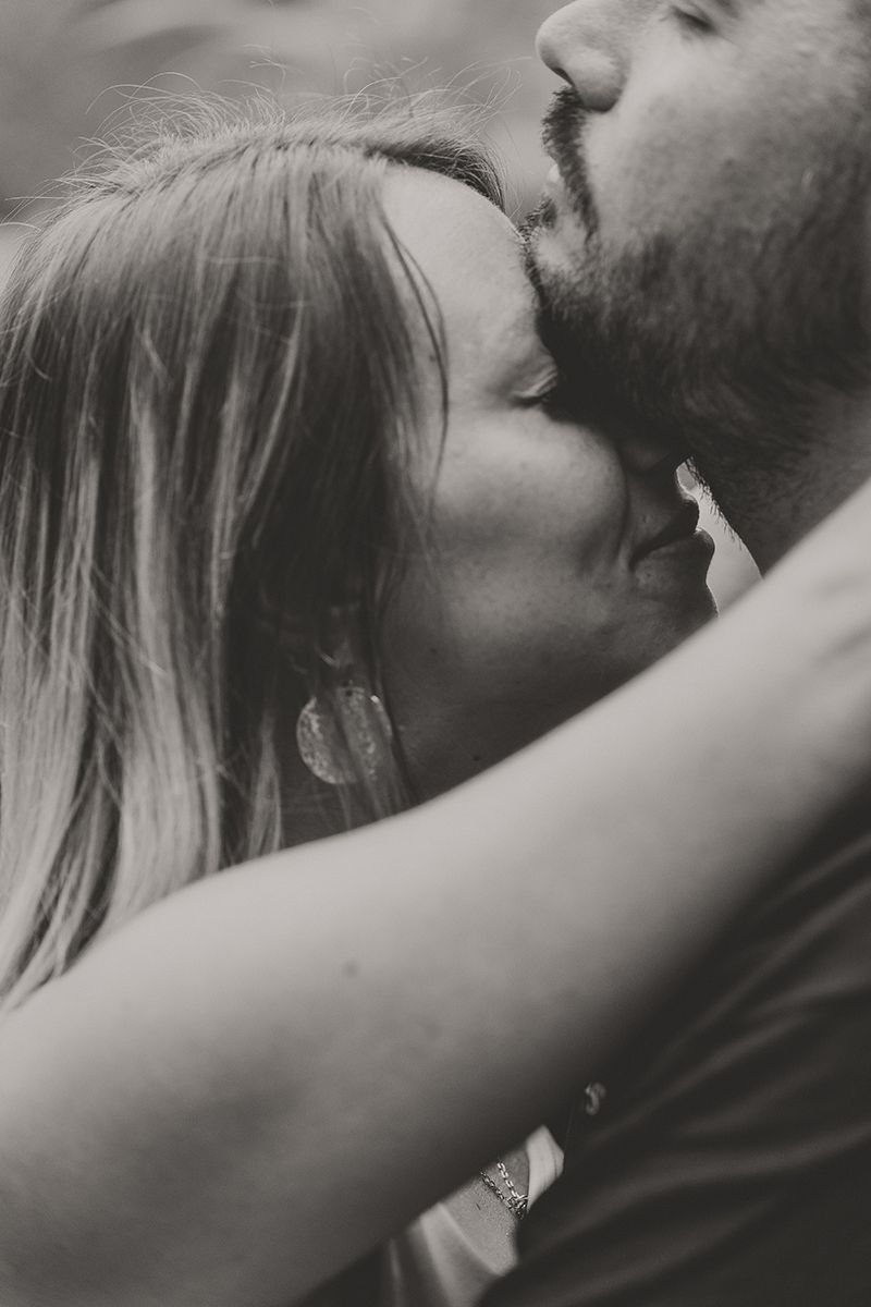 In a black and white close-up, a bearded man tenderly kisses a woman's forehead as she embraces him.