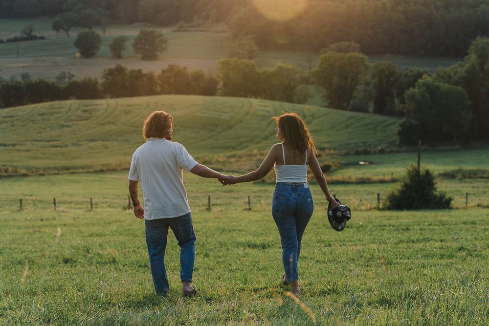 A couple holds hands and walks through a green countryside meadow with rolling hills and trees in warm evening light.