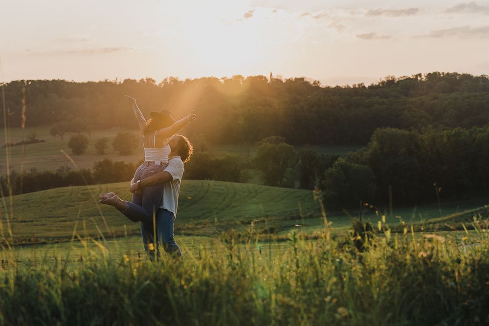 A man lifts his partner into the air in a joyful embrace in a grassy field as the setting sun glows over distant tree-lined hills.