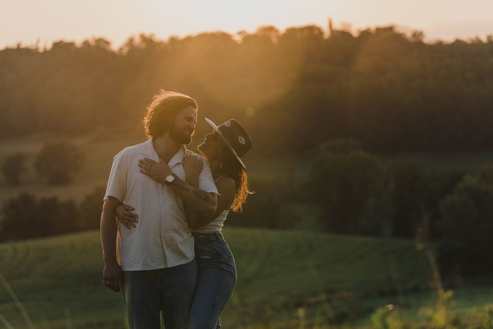 A couple embraces in a sunlit meadow at golden hour, the woman in a brimmed hat, with hazy rolling hills and trees behind them.