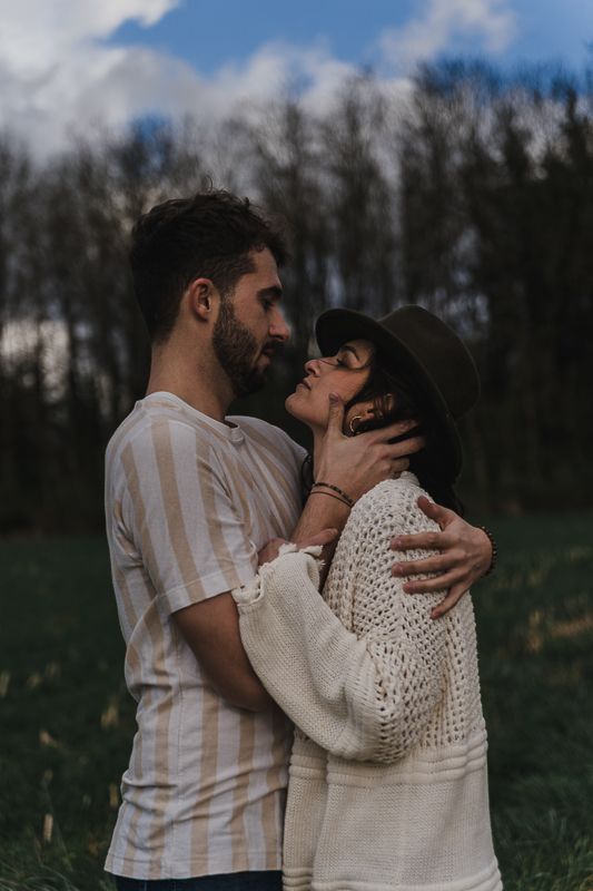 A bearded man in a striped shirt embraces a woman in a wide-brimmed hat and white knit cardigan in a grassy field at dusk.