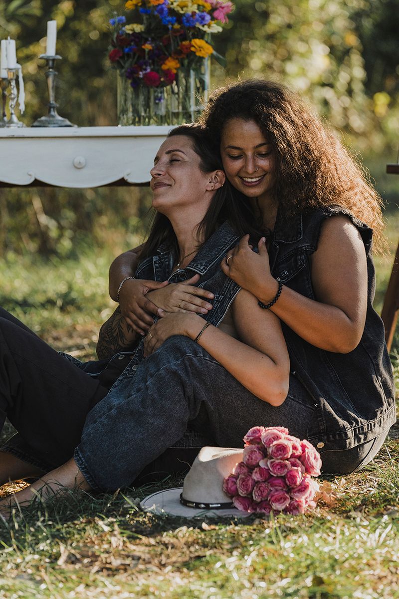 Two women in dark denim outfits sit close and embrace on the grass near a vintage table with flowers, candles and a pink bouquet.