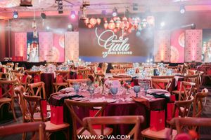 Elegant gala evening banquet hall with round tables set in red and gold, wooden chairs, and a glowing Gala Evening sign on a stage backdrop.