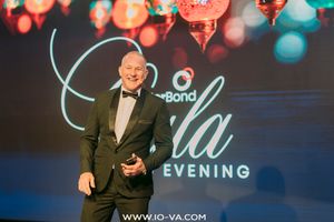 Man in a dark green velvet blazer and bow tie holding a microphone on stage at a gala evening event with glowing lanterns behind him.