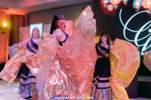 Three belly dancers in flowing dark skirts perform with shimmering golden iridescent wings on a stage at a festive indoor event.