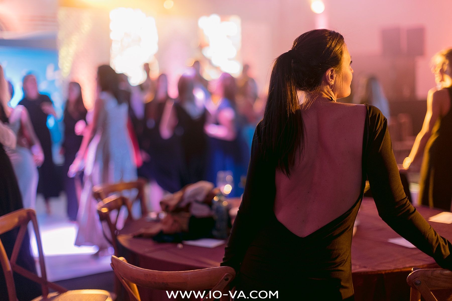 Woman with a ponytail stands beside a table at an elegant gala event while guests in formal evening wear mingle in the glowing background.