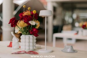 Red roses and yellow flowers in a white textured vase on a wooden table beside a modern cordless white lamp and a small ceramic rabbit.