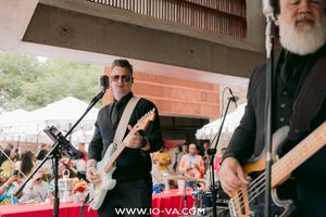 Two musicians play electric guitars and sing into microphones on an outdoor stage while a crowd gathers under white tents in the background.