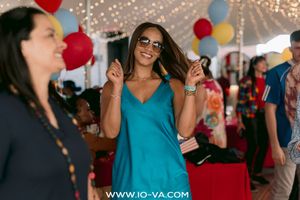 Woman in a teal dress and sunglasses dances at a festive party with colorful balloons and string lights while guests mingle behind her.
