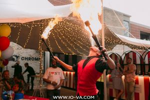 Fire performer in a red vest juggles two flaming torches at an outdoor event under string lights while guests watch.