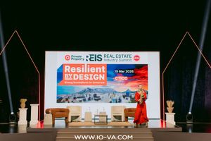 Speaker in a red dress stands on stage beside armchairs at the Resilient by Design EIS Real Estate Industry Summit presentation.