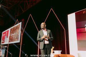 Speaker in a gray blazer presents on stage at a conference with glowing orange geometric lighting and large video screens behind him.