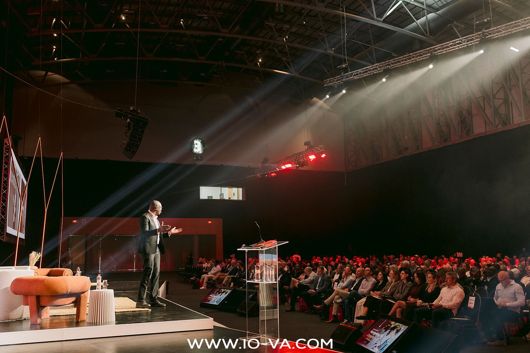 Speaker standing on a spotlit stage addressing a large seated audience in a dark conference hall with a podium and orange chair nearby.
