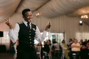 Smiling host in a vest with epaulettes gestures animatedly while speaking to seated guests in a draped event tent.