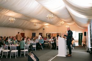 Speaker addresses seated guests from a podium inside a large white draped event marquee with chandeliers and round dining tables.