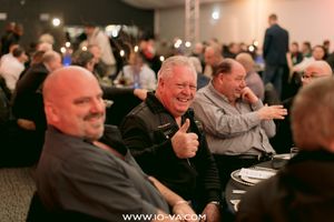 Smiling man in a black jacket gives a thumbs up while seated at a crowded banquet table at a dimly lit indoor event.