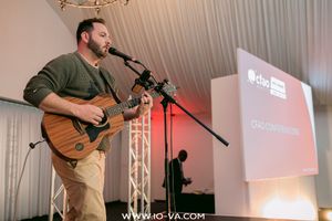 Bearded man in an olive sweater plays an acoustic guitar and sings into a microphone on stage beside a presentation screen at a live event.