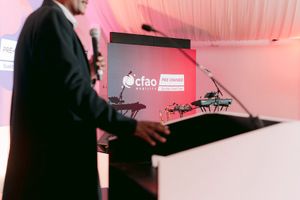 Speaker in a dark suit holds a microphone at a podium on stage, with a CFAO presentation screen glowing red in the background.