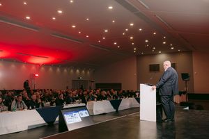 Speaker in a suit addresses a large audience seated at draped tables during a conference in a modern hall with red lighting.