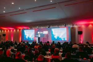 Speaker presenting on stage to a large seated audience at a conference, with illuminated screens and red ambient lighting in the hall.