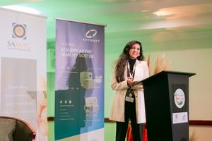 Woman in a white blazer speaks at a podium during a conference, with SAMAC and AI sorter banners displayed behind her.