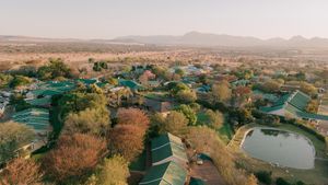 Aerial view of a safari lodge with green-roofed buildings, autumn-colored trees and a small pond amid savanna plains and distant hills.