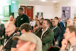 Audience members seated at a conference as a man in a green jacket holds a microphone to ask a question while others look on.