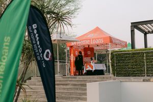 Person stands at an orange Home Loans promotional tent beside stone steps, with green and black Home Loans flags in the foreground.