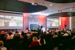 Audience seated in a modern conference auditorium watching a presenter on large screens during a business event with red curtain accents.