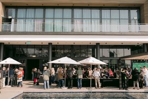 Crowd of people gathers under white patio umbrellas outside a modern glass-fronted building with an upper balcony, beside a reflecting pool.