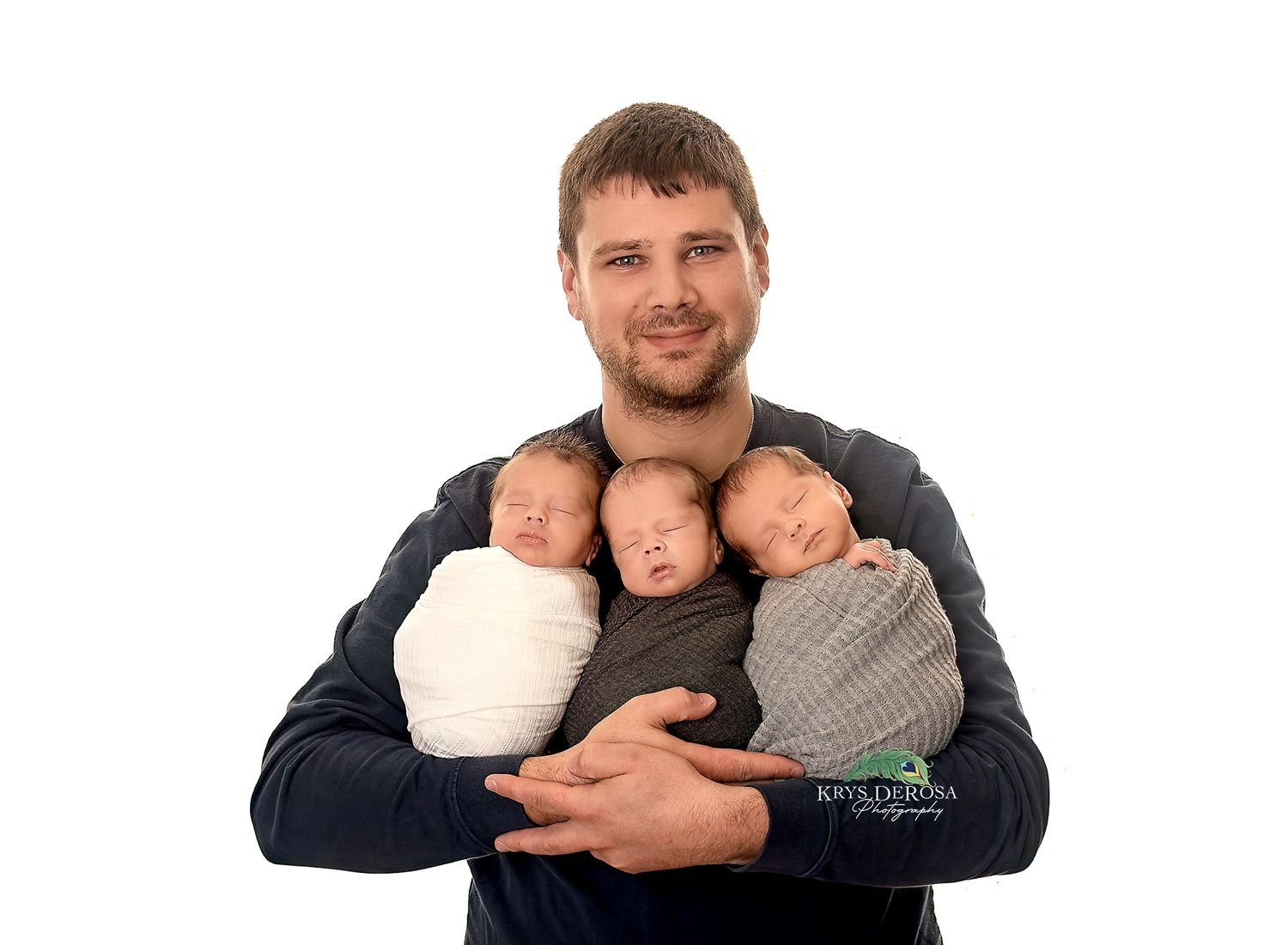 A smiling man in a navy shirt holds three sleeping newborn babies wrapped in white, beige, and gray swaddles.