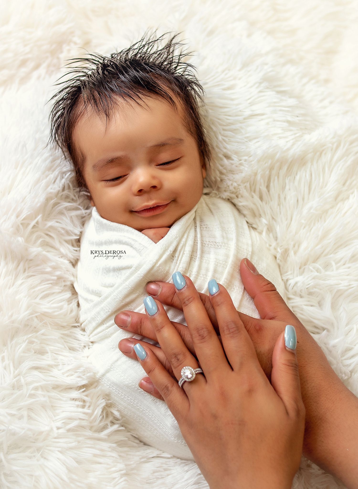 Newborn baby swaddled in white wrap sleeping peacefully on fluffy rug while mother's hands with blue nails and diamond ring rest gently.
