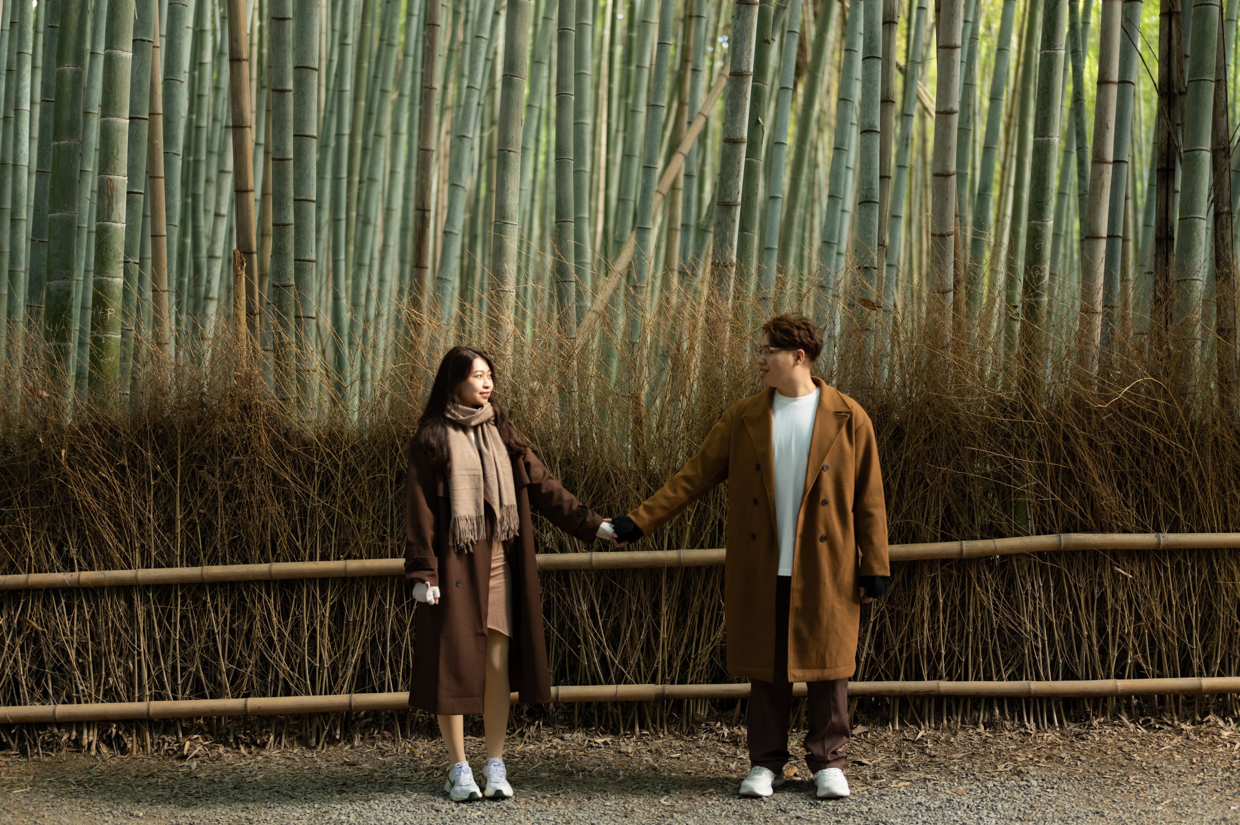 Couple in long brown coats holding hands while standing in front of a tall green bamboo grove with a wooden fence.