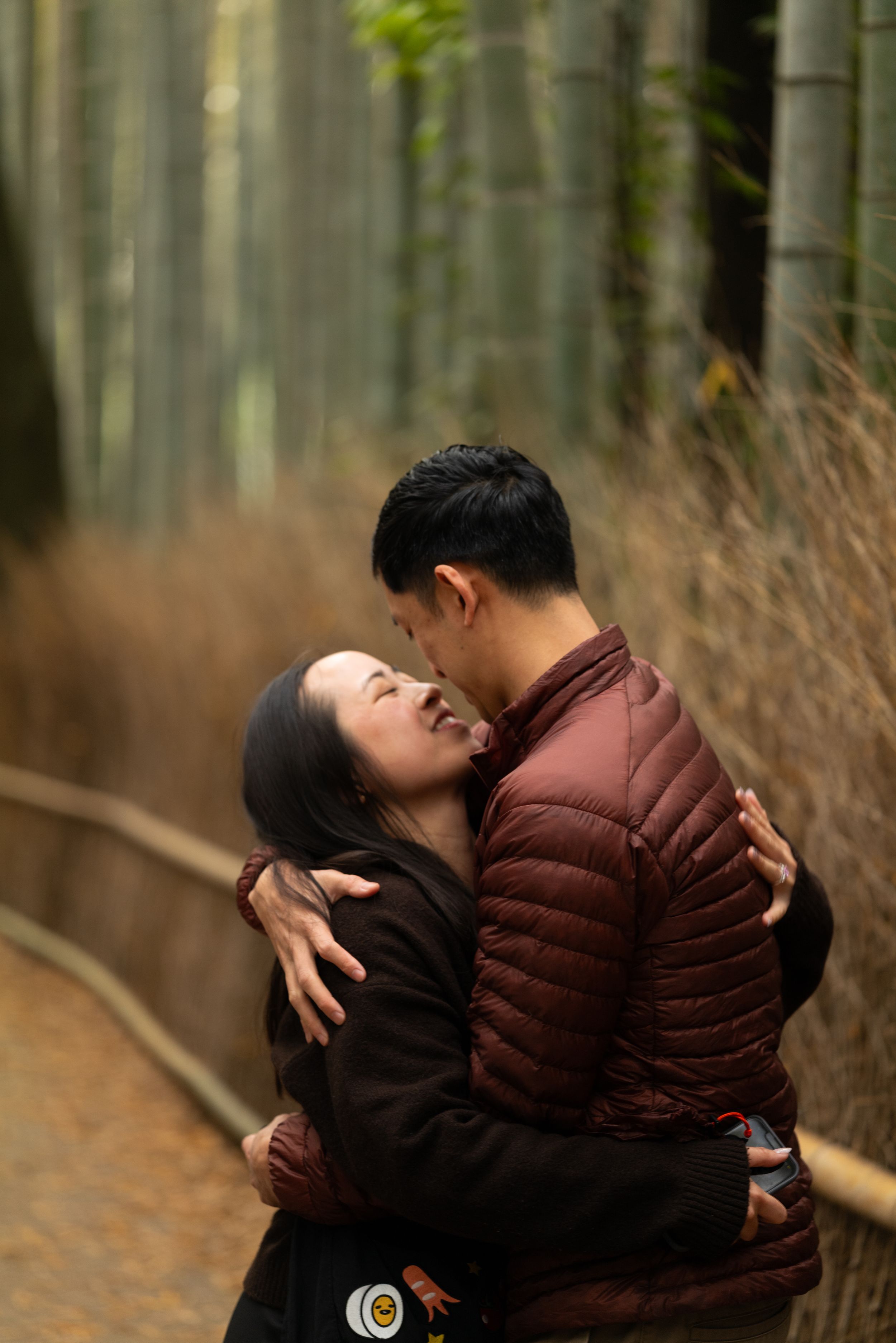 A couple embraces and laughs affectionately on a quiet path surrounded by tall bamboo trees in a peaceful forest.