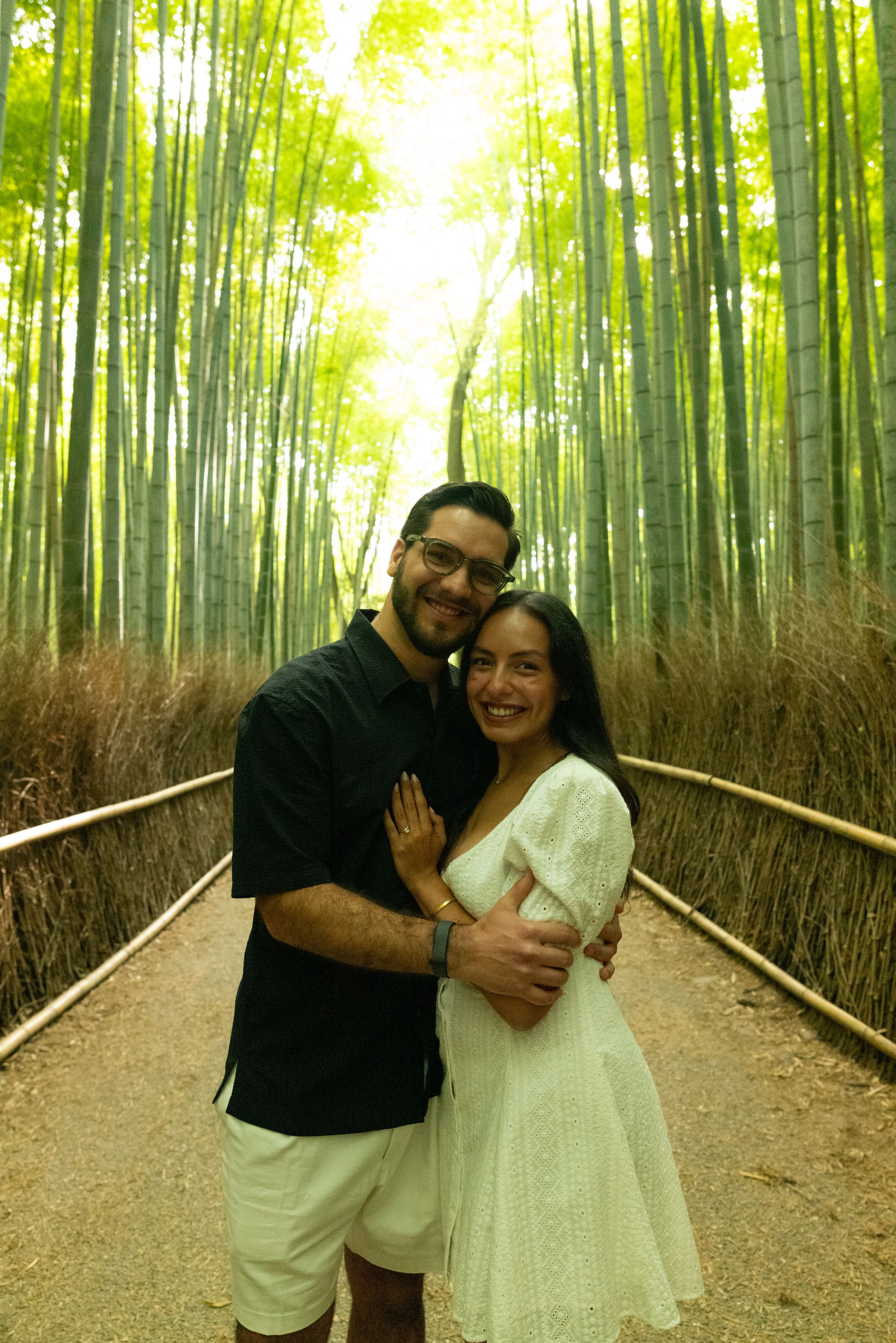 A smiling couple embraces on a dirt path lined with railings, surrounded by tall green bamboo trees glowing in warm sunlight.