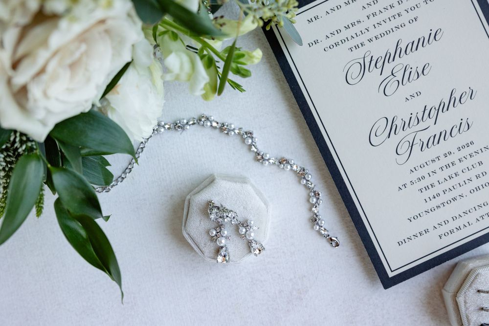 Wedding invitation with crystal drop earrings, diamond bracelet, and white roses on marble surface.
