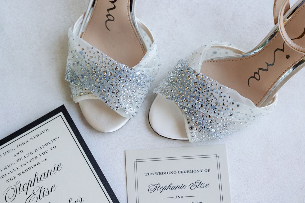 Sparkly crystal-embellished white wedding heels displayed beside elegant Stephanie Elise wedding ceremony invitations.