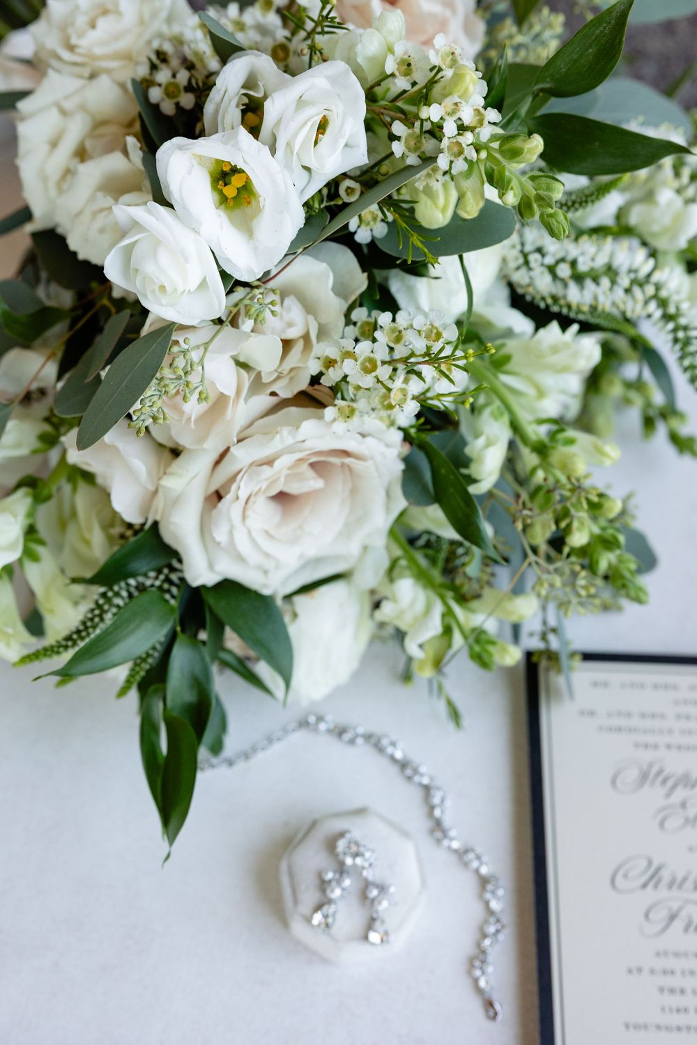 Elegant bridal flat lay featuring white roses, greenery bouquet, crystal earrings, bracelet, and wedding invitation on white surface.