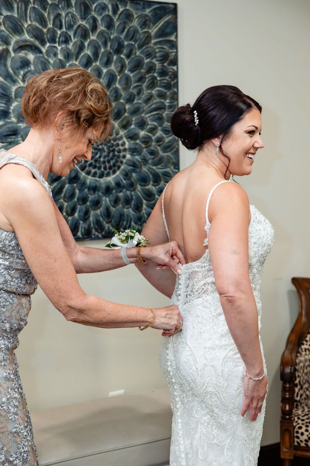 Mother helping bride button elegant backless beaded wedding gown while both smile joyfully before ceremony.