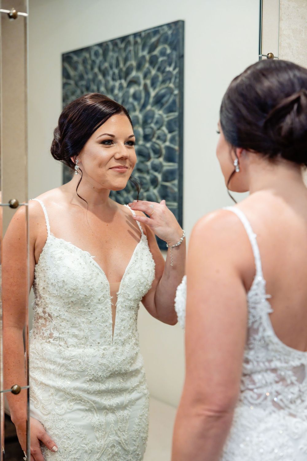 Bride in lace wedding gown admires her reflection in mirror while adjusting necklace before ceremony.